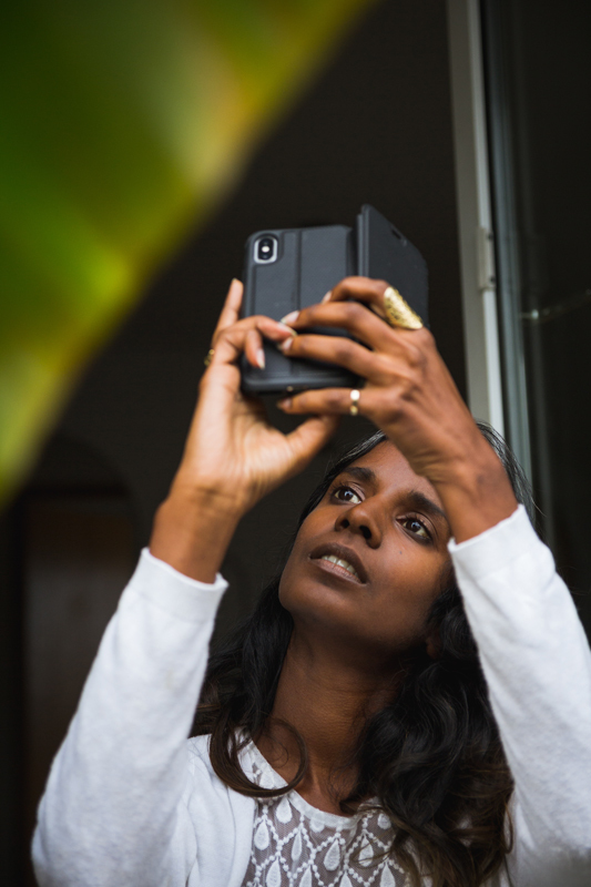 Low angle of focused ethnic female standing near glass balcony door and taking picture of plant for social media on mobile phone camera